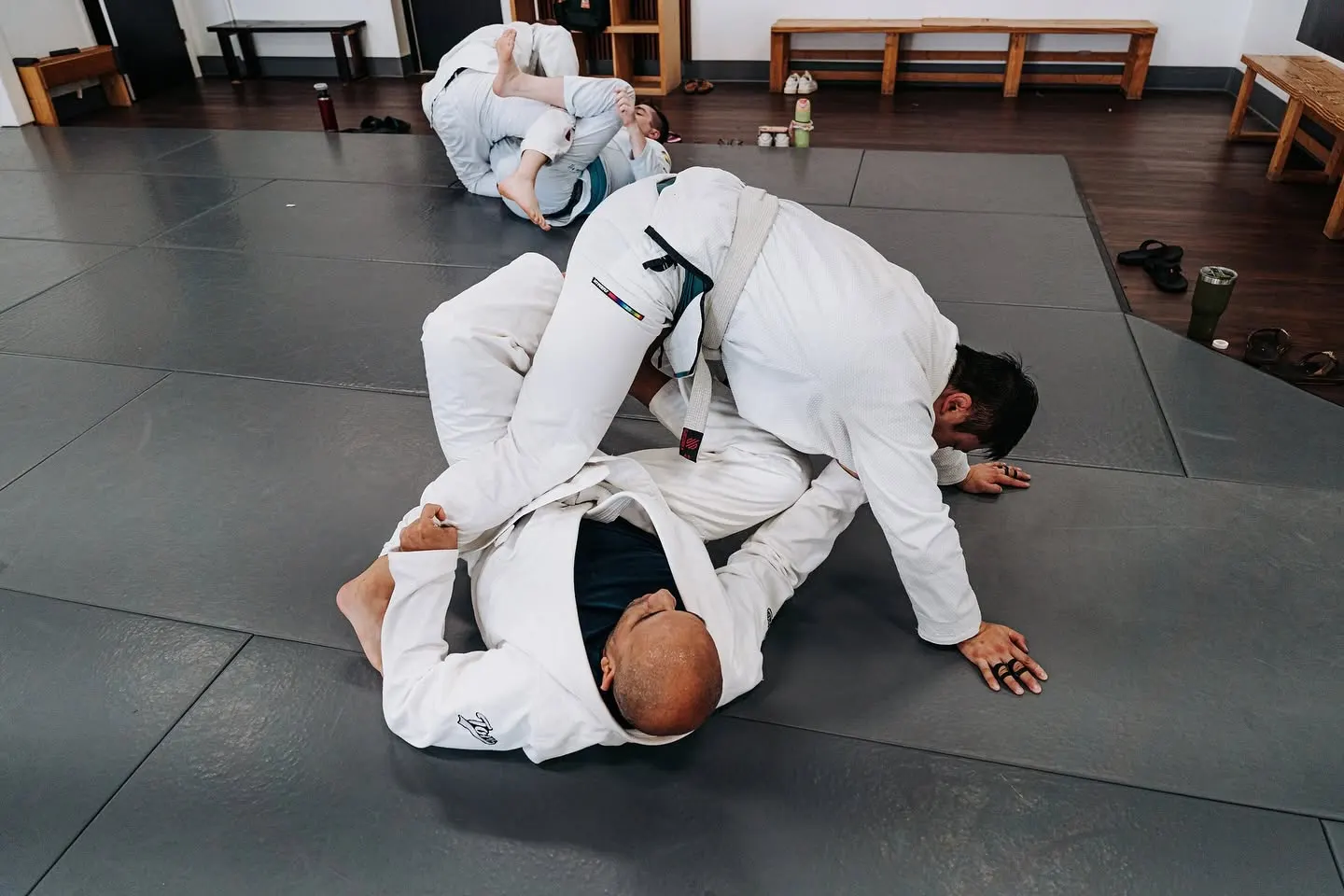 Two adults in white gis training a ground position on the mat during an adult Gi jiu-jitsu class